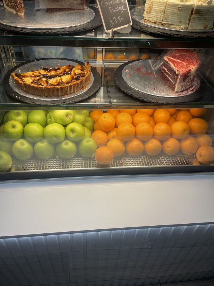 A chilled display case with an apple tart and a slice of layered cake above trays of apples and oranges