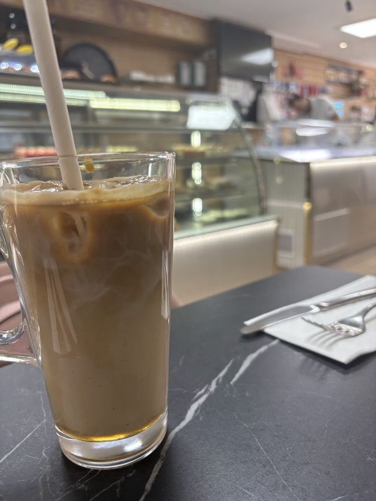A tall glass of iced coffee with a straw, the lit pastry counter blurred behind
